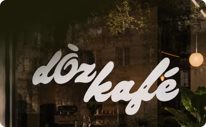 White cursive lettering spelling 'döz kafé' on a glass window with reflections of buildings and plants.