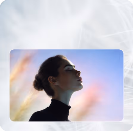 Side profile of a woman with hair in a bun wearing a black top, against a blurred blue sky background with soft pink and yellow light effects.